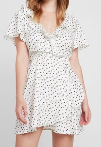 Valkoinen mekko mustilla pilkuilla, röyhelökaulus ja -hihat, solmittava vyötäröyksityiskohta ja kerroksellinen hame. Kevyt kangas, polven mittainen.