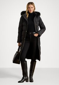 Čierna prešívaná bunda s kapucňou s kožušinovým lemom, predným zipsom, bočnými vreckami, v kombinácii s čiernym rolákovým svetrom, tmavými džínsy a čiernymi koženými čižmami do kolien.