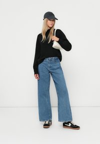 Svart genser, lyseblå vide jeans, svart og hvit sneakers, grå caps, holder en liten hvit håndveske, nøytral bakgrunn.