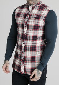 Koszula w kratę w czerwone, czarne i białe wzory. Długie granatowe rękawy, zapinana na guziki z haftowanym logo na piersi.