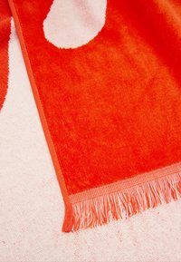 Miękki ręcznik z bawełny z wyrazistymi pomarańczowymi i białymi wzorami. Wykończony dekoracyjnym frędzlem wzdłuż jednej krawędzi.