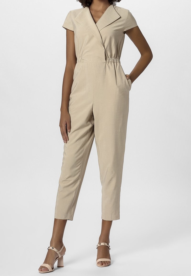 Femme portant une combinaison beige à manches courtes avec un large col et une taille élastique, associée à des sandales à talons blocs rose clair.