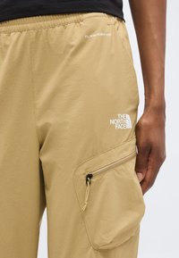 Μπεζ σορτς North Face με πλαϊνή τσέπη με φερμουάρ και λογότυπο FLASHDRY-XD, φοράει άτομο με σκούρο δέρμα και μαύρο πουκάμισο.