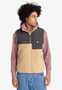 Fleece-Weste in Beige und Grau, mit Reißverschluss, Vordertaschen und einem markanten Patch. Getragen über einem rosa Hoodie mit klassischer Passform.