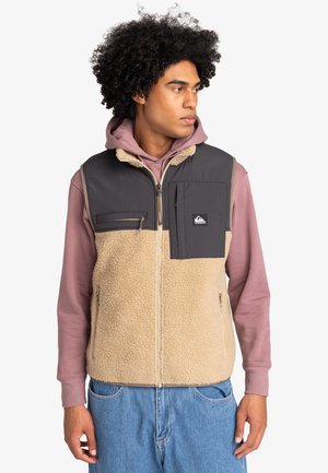 Fleece vest in tan and gray, featuring a zip closure, front pockets, and a distinctive patch. Worn over a pink hoodie with a classic fit.