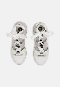 Chaussures de sport blanches avec une tige en mesh et cuir. Elles comportent des lacets épais blancs et une sangle grise. Design à bout ouvert avec une semelle texturée.