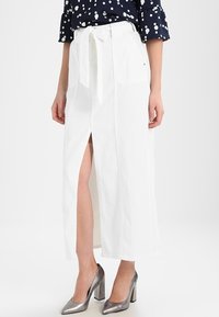 Mujer vestida con una falda midi blanca de cintura alta con abertura frontal y cinturón atado, combinada con zapatos de tacón plateados y una blusa azul marino con lunares.