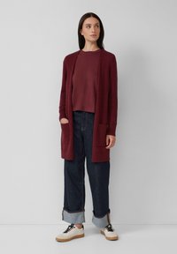 Femme portant un cardigan bordeaux et un haut, un jean large retroussé de couleur sombre et des baskets blanches, se tenant contre un fond clair.