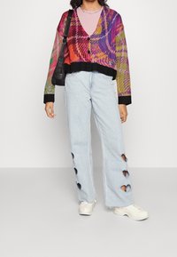 Osoba nosząca kolorowy sweter w wzory, jasnoróżową koszulę, jasnoniebieskie dżinsy z sercowymi wycięciami na nogach, białe tenisówki i czarną torbę na ramię.