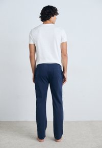 Marineblå bukser med en afslappet pasform, elastisk talje og baglomme, parret med en ensfarvet hvid t-shirt med korte ærmer. Gulvets tekstur er synlig.