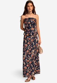 Ärmelloses, marineblaues, florales Maxikleid mit einem Peplum-Taille, das orange- und weiße Blumenmuster aufweist. Kombiniert mit einer beigen Clutch und braunen Absätzen.
