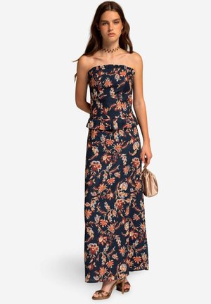 Mouwloze marineblauwe maxi-jurk met een peplum taille, voorzien van oranje en witte bloemenpatronen. Gecombineerd met een beige clutch en braune hakken.