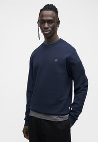 Μπλε σκούρα μπλούζα φούτερ με στρογγυλή λαιμόκοψη και μακριά μανίκια, με μικρό λευκό λογότυπο "II" στο στήθος, κατασκευασμένη από λείο, υφασμάτινο ύφασμα με υφή.