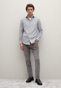 Chemise gris clair à boutons avec un motif à carreaux subtil, associée à un pantalon en velours côtelé beige et des mocassins noirs, posée devant un fond neutre.