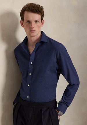 Jeune homme aux cheveux bouclés portant une chemise bleu foncé boutonnée et un pantalon noir, debout devant un mur beige uni.