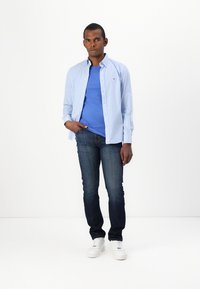 Camisa a cuadros azul claro sobre una camiseta azul, combinada con jeans oscuros y zapatillas blancas. Cuenta con cierre de botones y cuello.