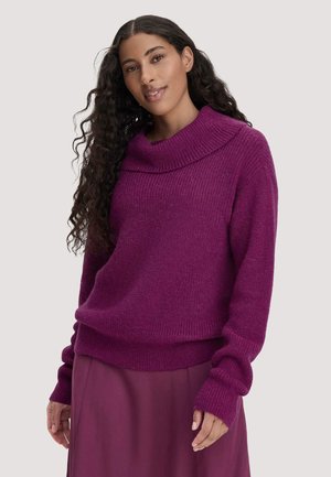 Woman with long curly hair wearing a purple knit cowl neck sweater and matching skirt, standing against a plain light background.