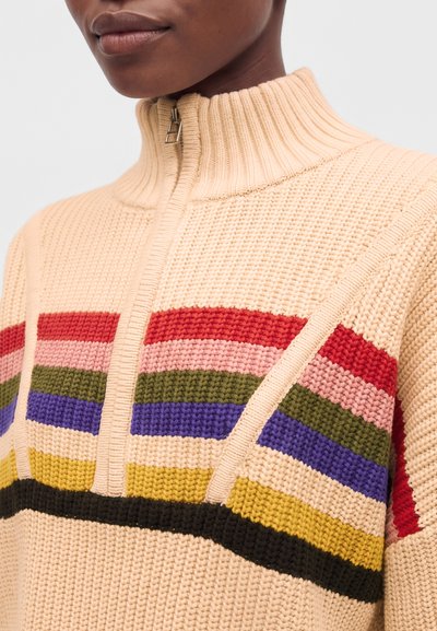 Gros plan d'une personne portant un pull en tricot beige avec un col zippé et des rayures horizontales rouges, roses, vertes, bleues, jaunes et noires.