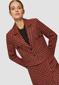 Marynarka w czerwono-czarny wzór w houndstooth z kołnierzem, dwoma przednimi kieszeniami i czarnymi guzami, wykonana z teksturowanej tkaniny.