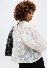 Γυναίκα με σγουρά μαλλιά, που φοράει διαφανές λευκό μπλουζάκι από δαντέλα με λουλούδια, κρατώντας μια μαύρη, υφασμάτινη τσάντα στον ώμο της, σε πλάγια όψη.