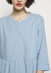 Blå och vit rutig skjorta med rund halsringning, knappstängning, trekvartslånga ärmar och en volangkjol. Tillverkad av bomull.