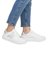 Baskets en cuir blanc avec un bout rond, des lacets plats, des accents argentés et une semelle en caoutchouc texturée pour l'adhérence. Jeans avec un ourlet retroussé.