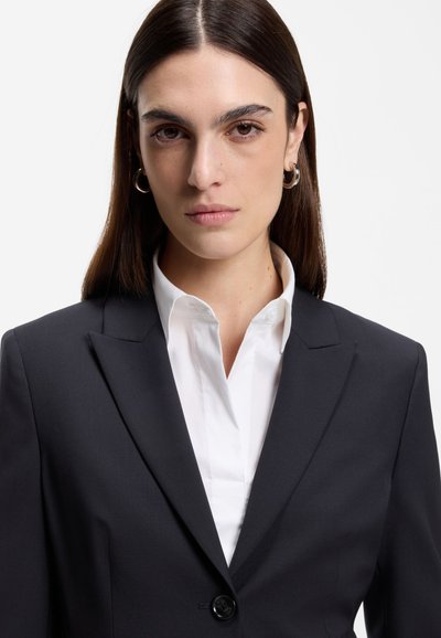 Femme aux cheveux raides et foncés portant des boucles d'oreilles argentées en forme de créoles, une chemise blanche à col et un blazer noir, faisant face à l'avant avec une expression neutre.