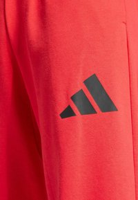 Červené sportovní kalhoty vyrobené z měkké látky s velkým černým logem Adidas na levé nohavici.