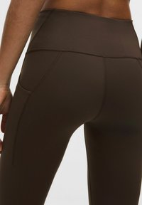 Lähedalne vaade isikule, kes kannab kõrge vöökohtadega tumepruune retuuse, millel on nähtavad küljetaskud ja õmbluse detailid.