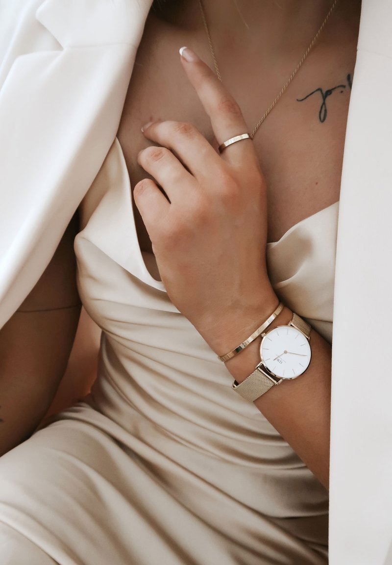 Gold mesh wristwatch with a white face, worn alongside a thin gold bracelet. The hand rests on a satin light-colored dress.