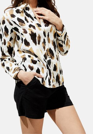 Femme portant une chemise blanche boutonnée avec un imprimé animal marron et noir et un short noir, une main dans la poche et l'autre près du col.