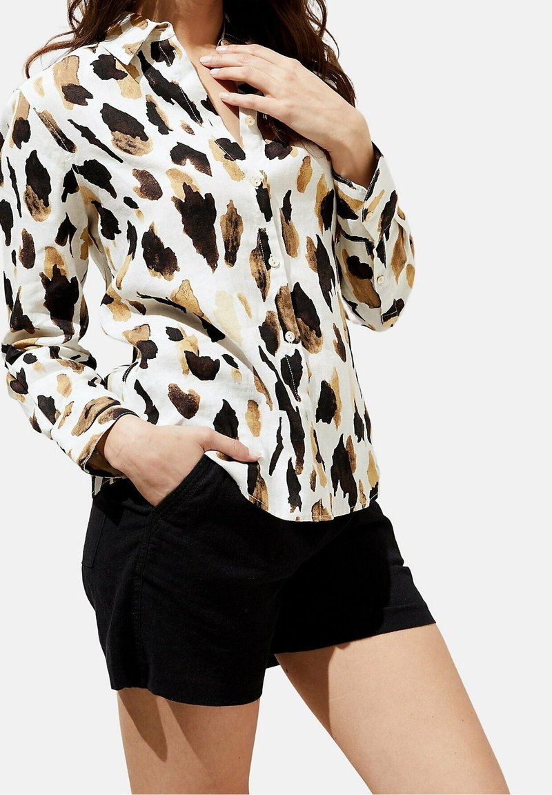 Femme portant une chemise blanche boutonnée avec un imprimé animal marron et noir et un short noir, une main dans la poche et l'autre près du col.
