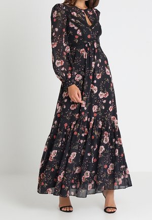 Femme portant une robe maxi noire à manches longues avec des fleurs roses et un ourlet à volants, associée à des talons ouverts noirs.