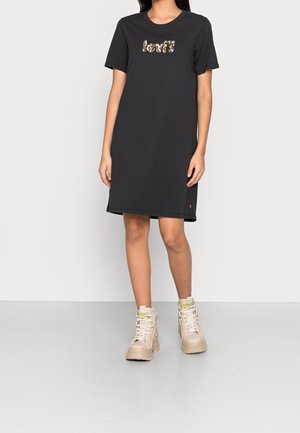 Kvinde iført en knælængde sort Levi's kjole med korte ærmer og blomsterlogo, beige høje chunky sneakers, stående mod en ensfarvet baggrund.