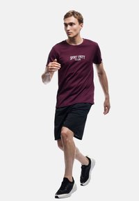 Camiseta deportiva marrón con el texto "CLUB DE UNIDAD DEPORTIVA", combinada con pantalones cortos negros y zapatillas negras. Modelo en movimiento, fondo neutro.