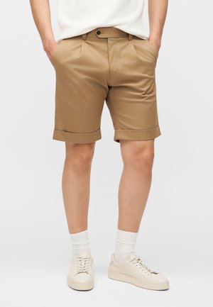 Άνδρας που φοράει μπεζ σορτς με γυρισμένη άκρη, λευκά αθλητικά παπούτσια και λευκές κάλτσες, στέκεται με τα χέρια στις τσέπες μπροστά σε απλό φόντο.