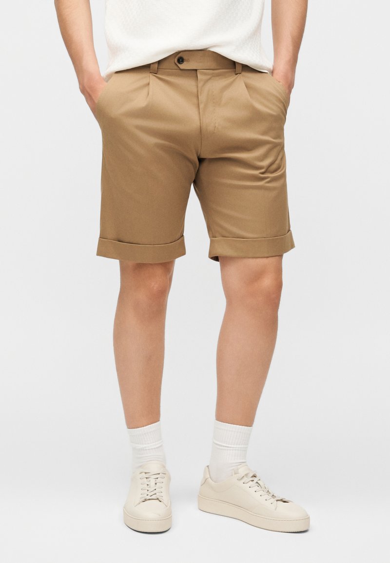 Άνδρας που φοράει μπεζ σορτς με γυρισμένη άκρη, λευκά αθλητικά παπούτσια και λευκές κάλτσες, στέκεται με τα χέρια στις τσέπες μπροστά σε απλό φόντο.