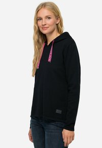 Felpa nera con cappuccio e lacci rosa, materiale testurizzato, vestibilità comoda e un'etichetta logo in basso a sinistra. Indossata con jeans in denim scuri.