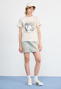 Tricou grafic bej cu logo adidas, fustă din denim cu un spălare în gradient, șapcă bej, șosete albe și adidași albi cu dungi maronii.