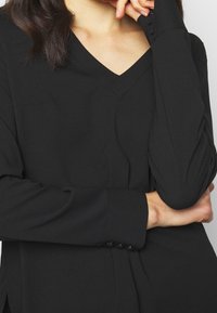 Blusa negra de cuello en pico con mangas largas y puños abotonados, textura suave y diseño minimalista, bolsillo parcialmente visible en la parte izquierda.