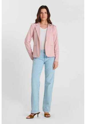 Vrouw staand, gekleed in een lichtroze blazer, wit shirt, lichtblauwe spijkerbroek en bruine sandalen met hak tegen een effen lichte achtergrond.