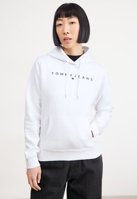 Felpa bianca con cappuccio caratterizzata dal logo "TOMMY JEANS" in nero, tasca frontale e polsini a coste. Indossata sopra una maglietta nera e abbinata a pantaloni scuri.