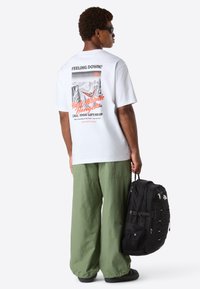 Muž v slunečních brýlích, bílém tričku s grafikou, širokých zelených kalhotách, držící černý batoh, stojící bokem na jednobarevném bílém pozadí.