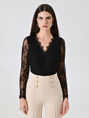 Zwarte kant bodysuit met een golvende halslijn en lange mouwen, gecombineerd met hooggerokte beige broeken met gouden knoopaccenten.