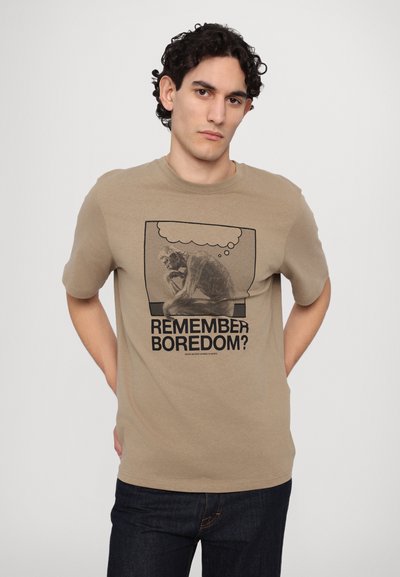 T-shirt en coton beige avec un motif noir représentant une figure et le texte "TE SOUVIENS-TU DE L'ENNUI ?" en caractères gras. Design à manches courtes et col rond.