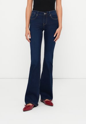 Donna vestita con jeans a zampa d'elefante blu scuro, top nero e ballerine rosse a punta, in piedi su uno sfondo bianco uniforme.