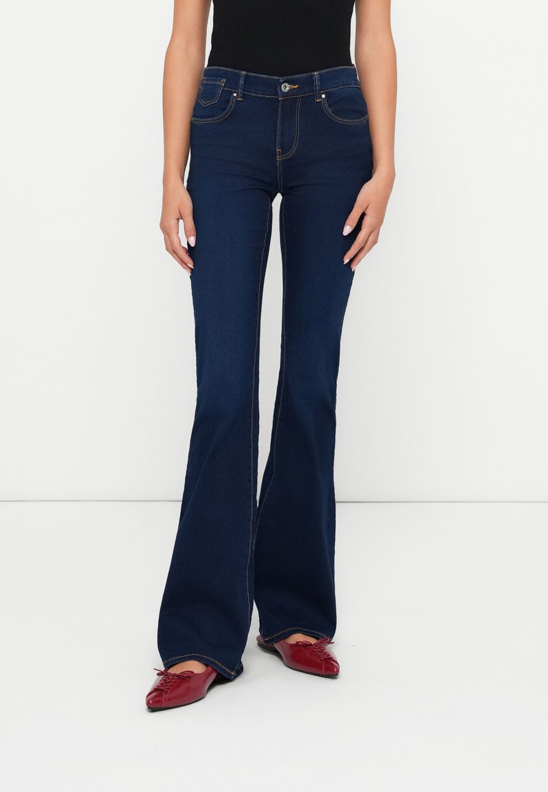 Mujer vistiendo jeans acampanados oscuros, top negro y zapatos planos rojos de punta, de pie contra un fondo blanco liso.