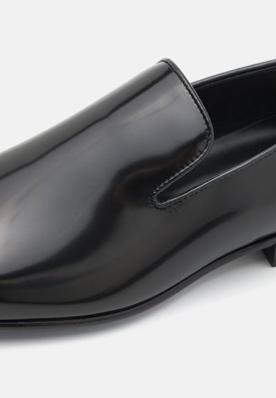 Sorte læderslips-on sko med en glat, shiny finish, rund tå og minimal syning. Lav blokhæl og ingen synlig hardware.