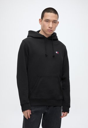 Młody mężczyzna w czarnej bluzie z kapturem z małym czerwonym, białym i niebieskim logo na piersi oraz w ciemnych jeansach, stojący na białym tle.