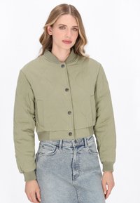 O femeie îmbrăcată cu o jachetă bomber matlasată de culoare verde deschis, cu nasturi metallici, și o fustă din denim de culoare albastră, cu talie înaltă, pe un fundal alb.